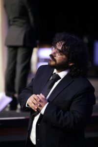 Caleb, a Latino man, looking away from the camera and slightly up, wearing a black dinner jacket, black tie, and white button down shirt. He is wearing metal framed glasses and has curly shoulder length hair. He has a mustache and beard as well. There is a blurred out background with someone in black dress pants standing on a stage behind him.