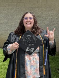 pronouns: she/her image description: Mallory, a white woman, is standing outside in daylight, smiling widely at the camera. She has shoulder-length, wavy, brown hair and is wearing round, wire-rimmed glasses. She is dressed in a black graduation gown with a light pink, blue, and white floral dress visible underneath. She has a golden cord and blue and orange cord draped around her neck. She is standing on green grass in front of a light tan brick wall. In her left hand, she holds up a decorated black graduation cap. The cap features silver outlines of hands in American Sign Language, signing “PAH” with the word spelled out underneath. It includes two hands signing the word “Interpreter” at the bottom. With her right hand, she is holding up the “I love you” sign in American Sign Language with a blue and white tassel hanging from her thumb.