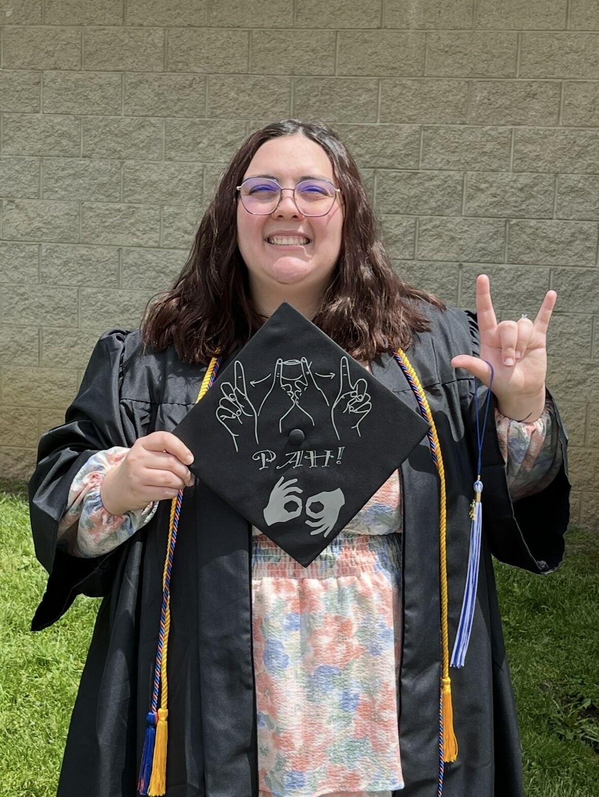pronouns: she/her image description: Mallory, a white woman, is standing outside in daylight, smiling widely at the camera. She has shoulder-length, wavy, brown hair and is wearing round, wire-rimmed glasses. She is dressed in a black graduation gown with a light pink, blue, and white floral dress visible underneath. She has a golden cord and blue and orange cord draped around her neck. She is standing on green grass in front of a light tan brick wall. In her left hand, she holds up a decorated black graduation cap. The cap features silver outlines of hands in American Sign Language, signing “PAH” with the word spelled out underneath. It includes two hands signing the word “Interpreter” at the bottom. With her right hand, she is holding up the “I love you” sign in American Sign Language with a blue and white tassel hanging from her thumb.