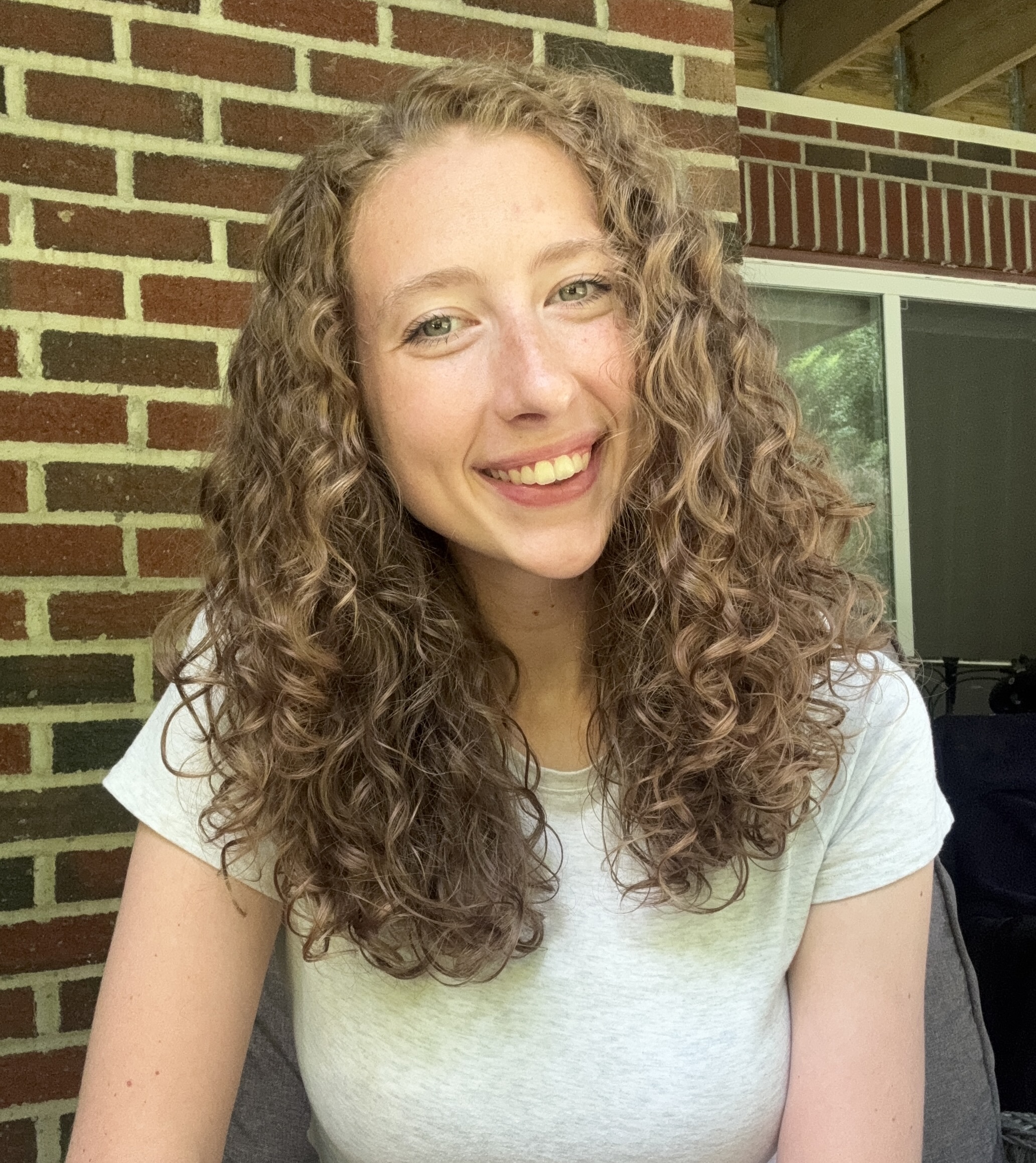 Rachel, a white woman with honey-colored curly hair and light eyes, smiles warmly at the camera. She is sitting in front of a brick wall and wearing a light grey shirt.