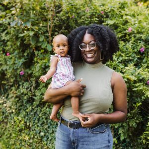 Shannon, a Black woman stands in front of greenery holding her young daughter as the both gaze toward the camera. They’re both wearing summer attire.
